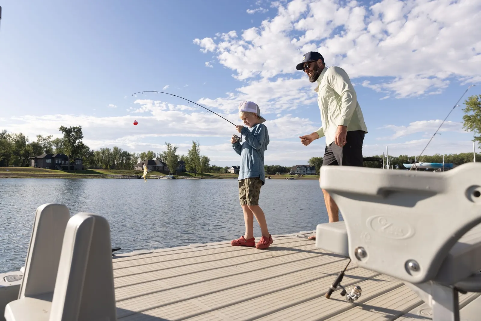 A father watching his young son play a bent fishing rod on a tan EZ Dock floating deck, lake houses along the far bank