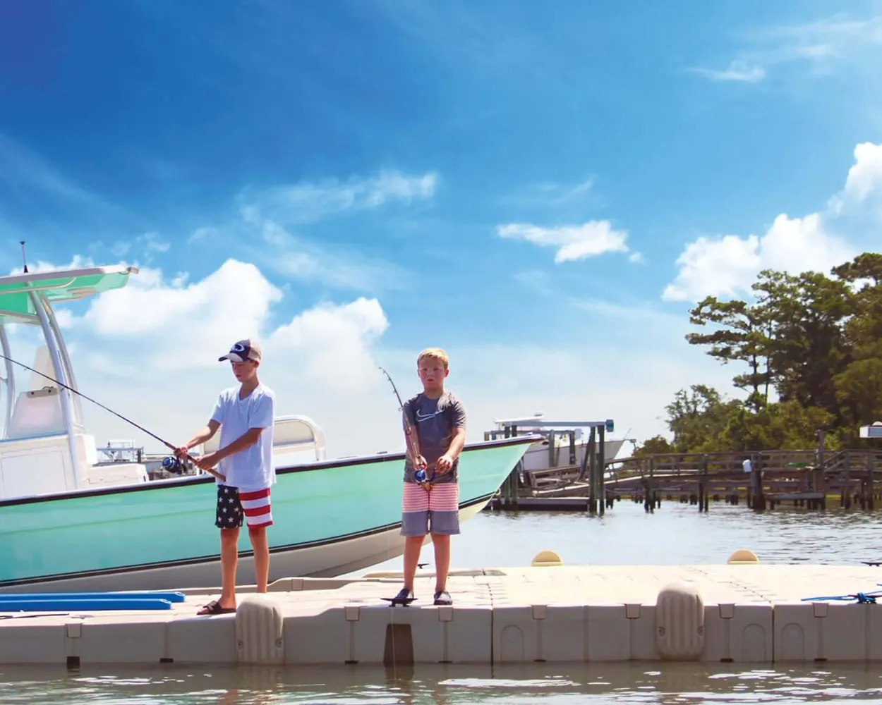 Two boys fishing side by side off a tan EZ Dock floating platform on a bright summer day, a boat tied up alongside