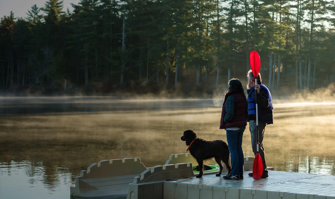A couple and their black dog standing on a tan EZ Dock floating dock at misty dawn, the man holding a paddle