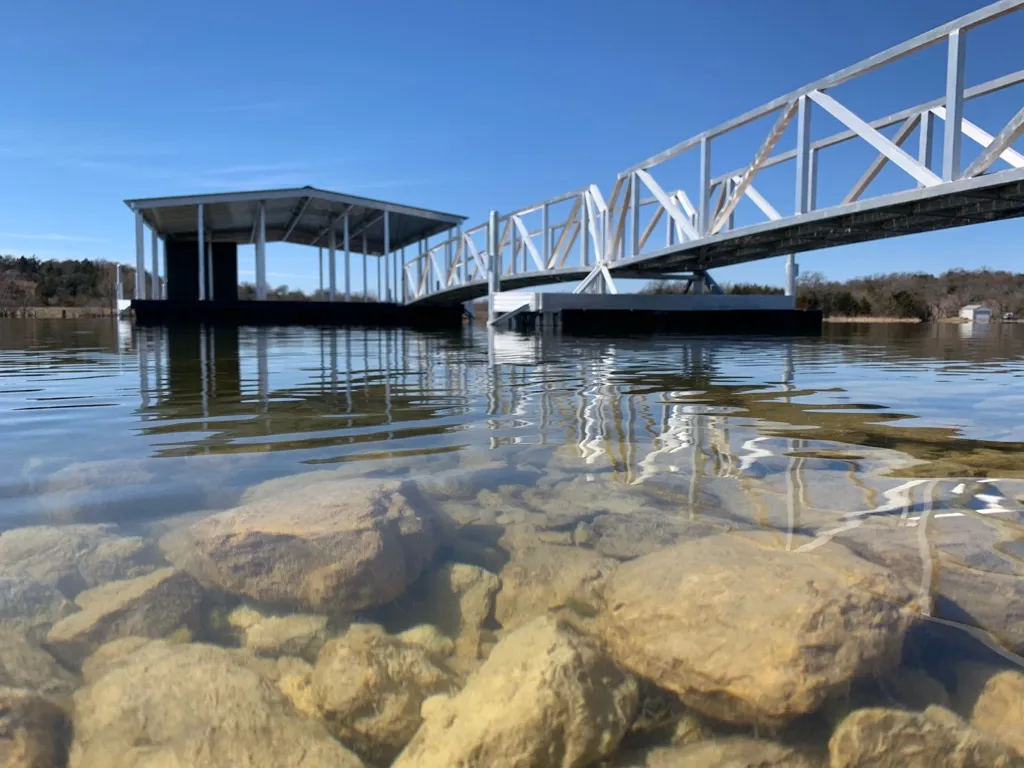 A finished covered dock and aluminum walkway seen low from the water, with rocks visible through the clear shallows under a blue sky.
