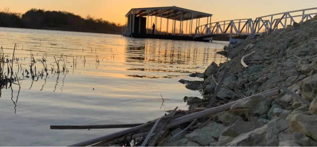 A covered dock silhouetted against a warm sunset, seen from a rocky bank at the water's edge.