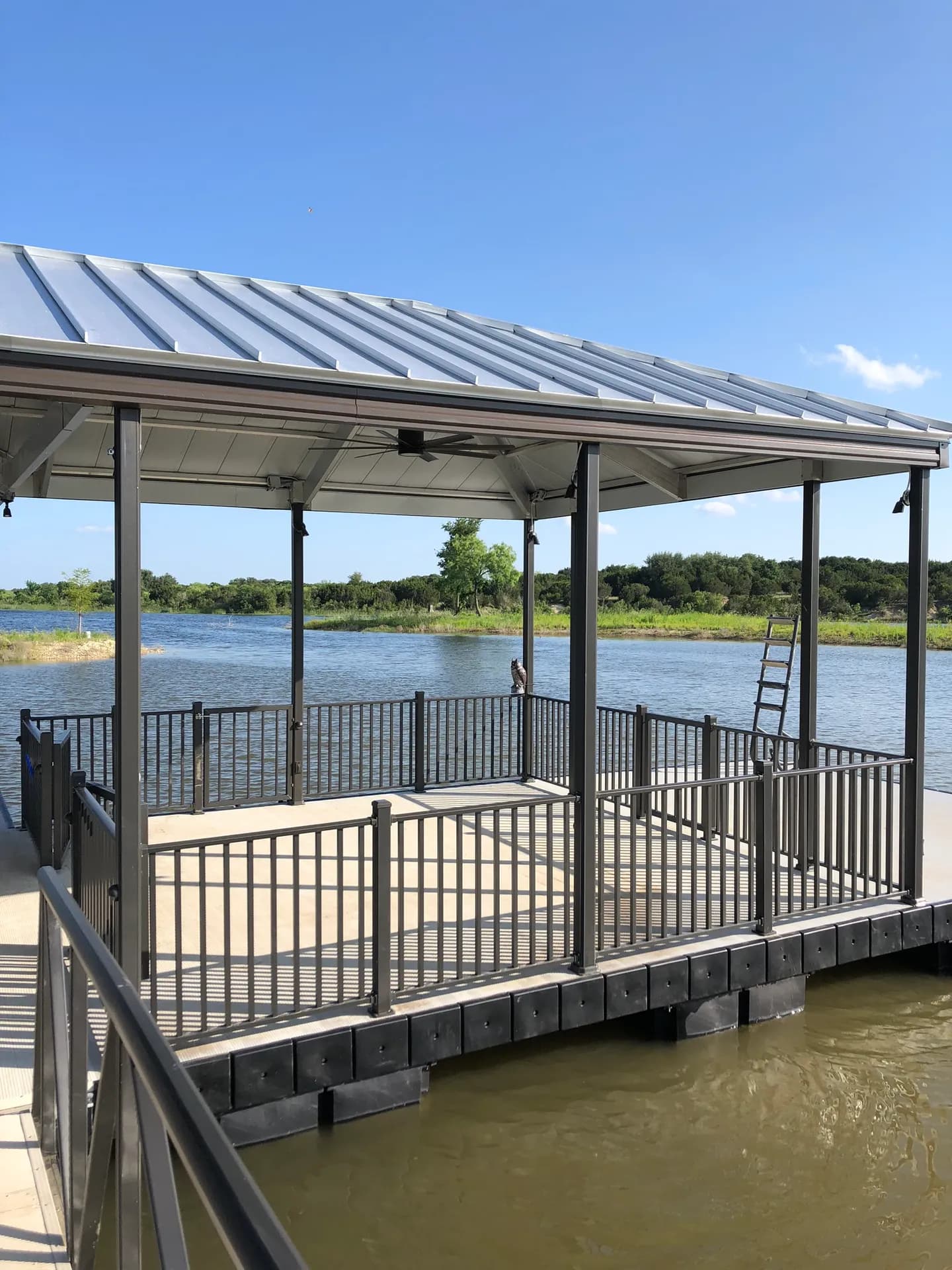 Covered aluminum dock with a metal roof, perimeter railing, and a swim ladder under a blue sky.