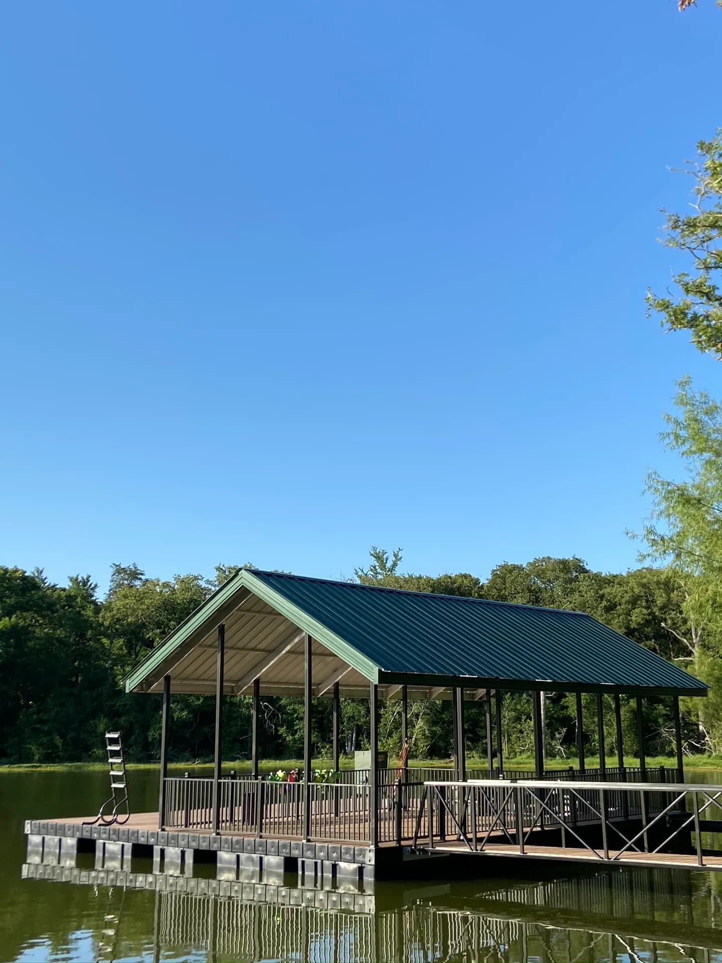 Covered dock with a green metal gable roof and composite deck, a swim ladder, and an aluminum walkway, under a clear blue sky.