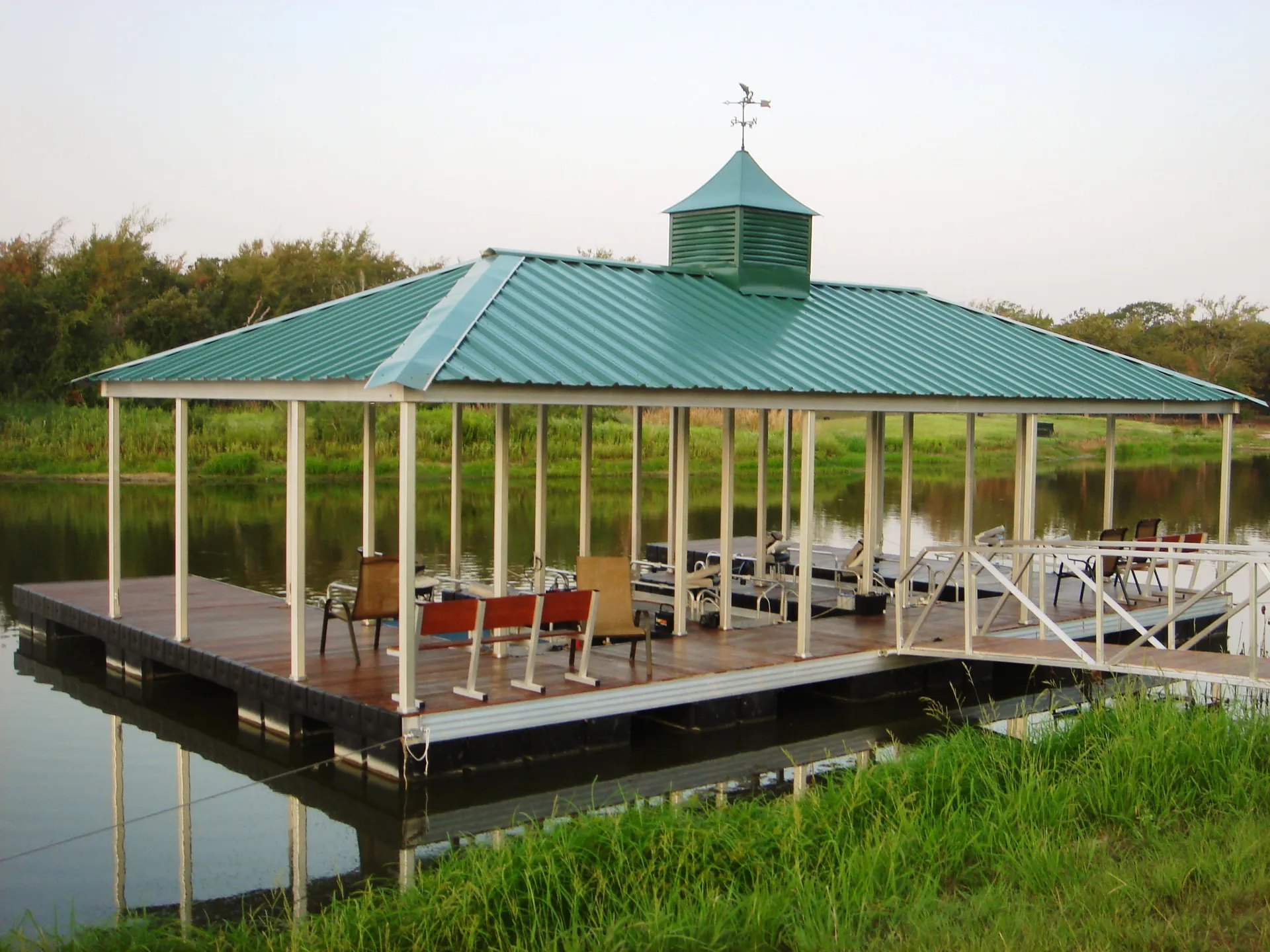 A covered aluminum dock with a green hip roof and a cupola, composite decking, and a swim ladder under a bright sky.