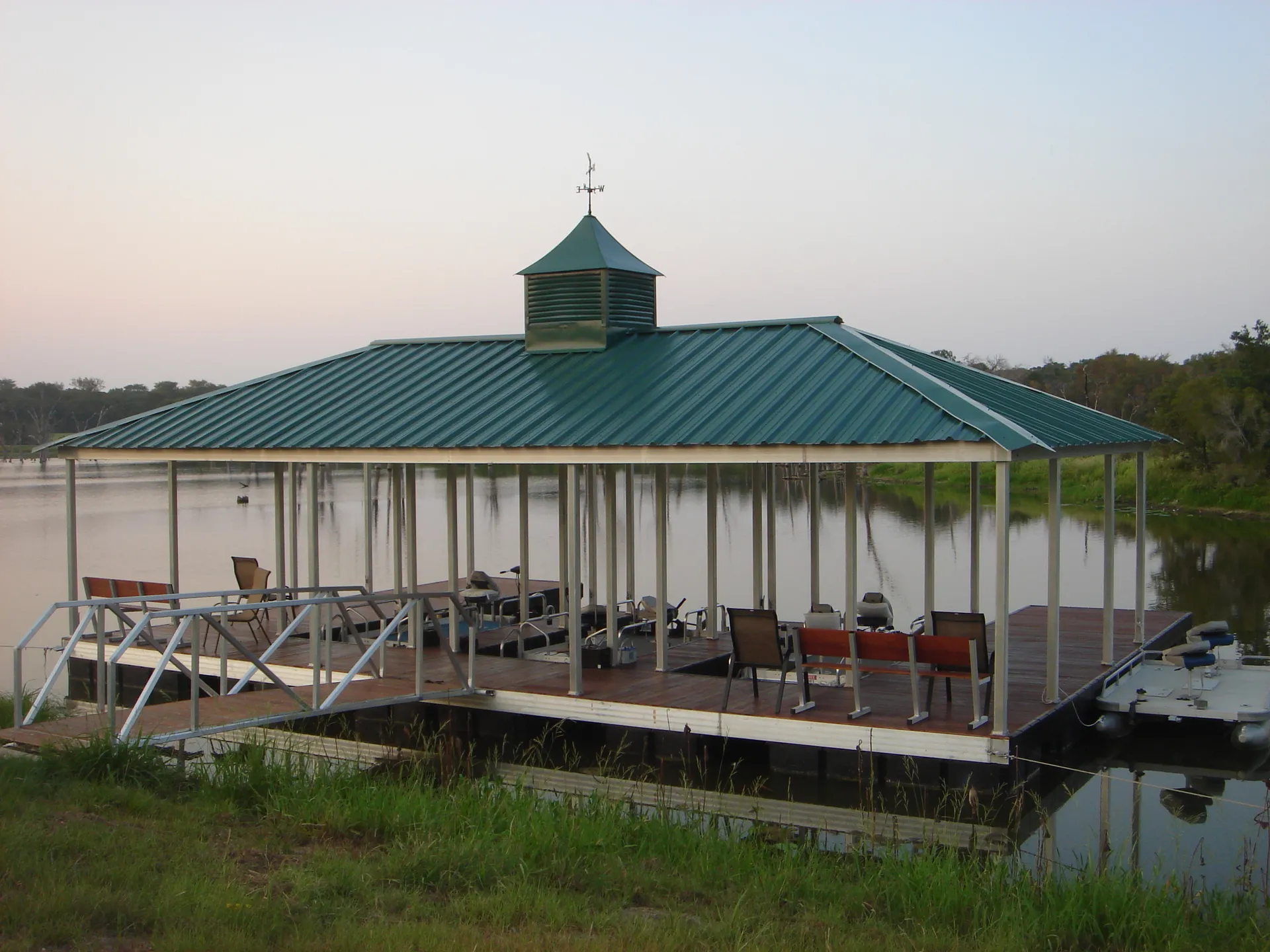 A covered aluminum dock with a hip roof and a cupola at dusk, its lights reflecting on still water.