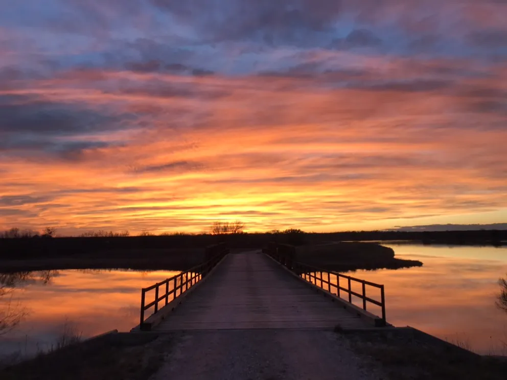 A wooden walkway leading straight out over calm water beneath a vivid orange-and-purple sunset.
