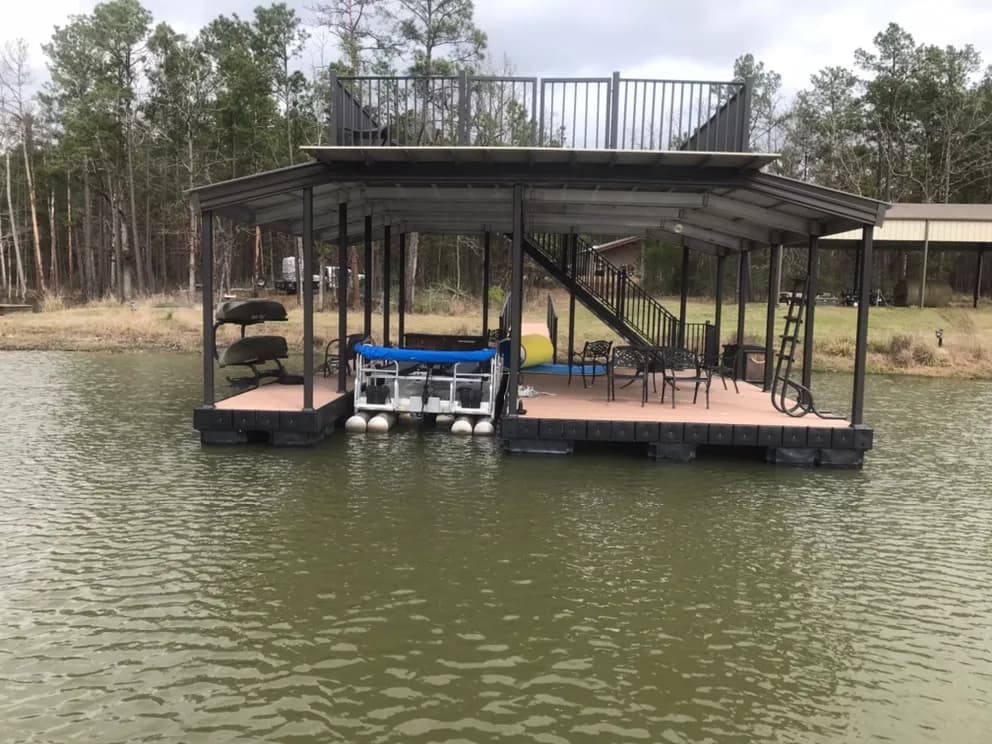Two-story covered aluminum dock viewed from the water, with a railed upper sun deck, patio seating, and a boat lift.