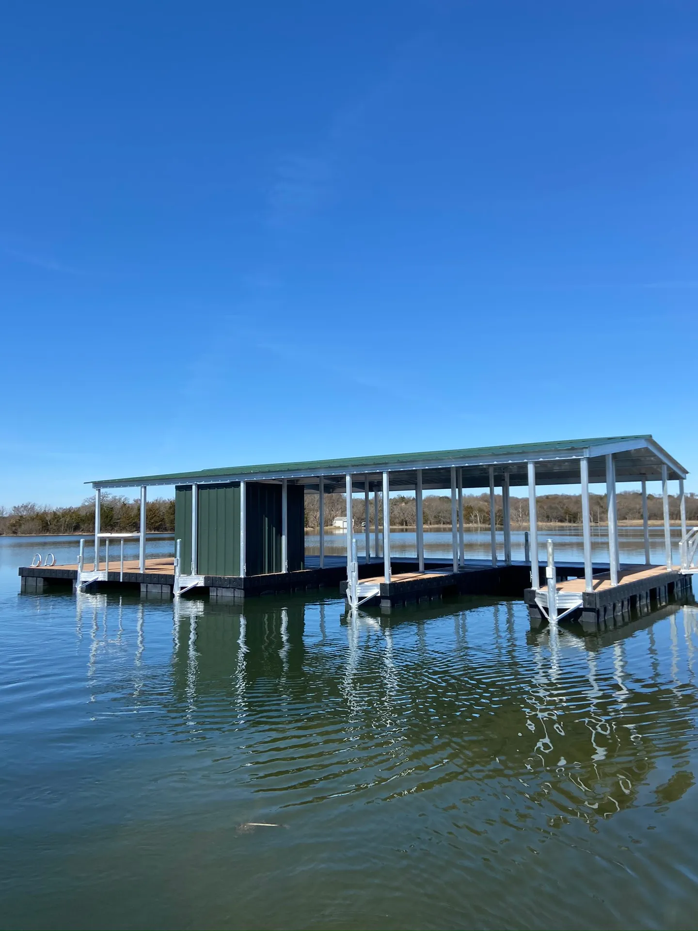 Long covered dock with white posts, a green metal roof, and multiple boat slips on open water under a clear sky.