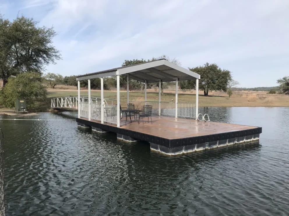 Sunlit covered dock with white posts and a gabled roof, a patio table and chairs on composite decking, and a railed walkway to the bank.
