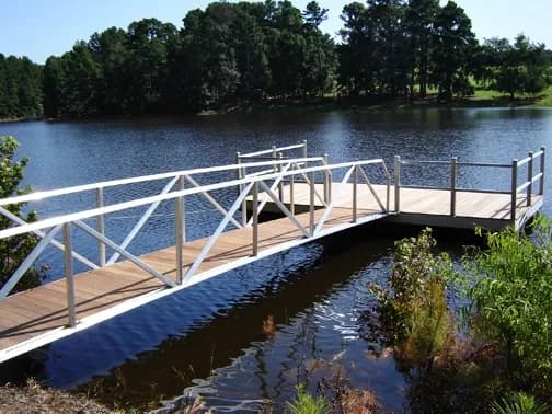 White aluminum truss walkway with railings leading to a composite-decked dock over a wooded pond.